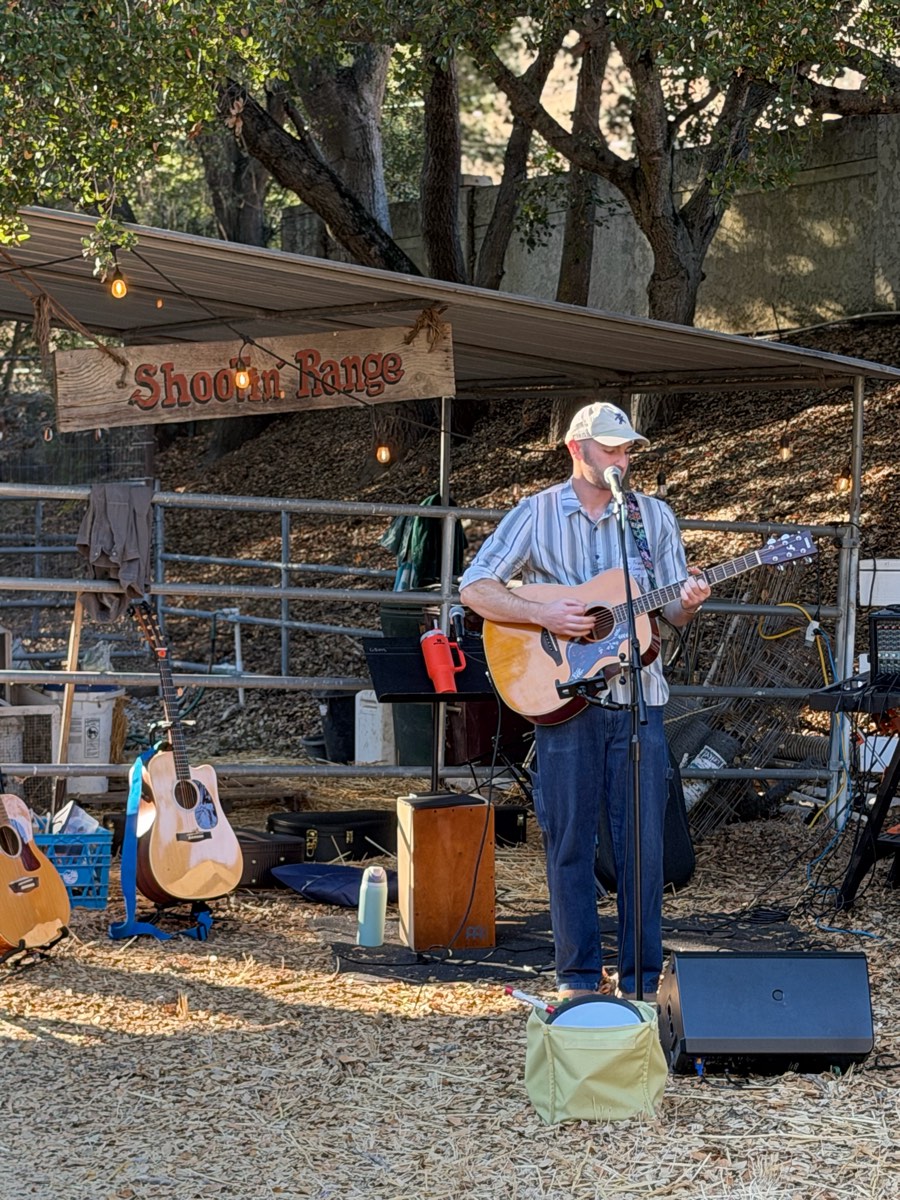 Playing acoustic guitar and singing solo at an outdoor show under a Shootin' Range sign